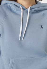 Sudadera azul clara con capucha, cordones blancos y un pequeño logo marinero. Fabricada con tejido suave; cuenta con un bolsillo frontal.