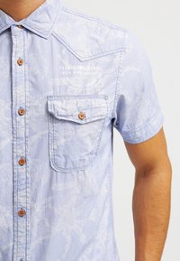 Vaaleansininen lyhythihainen paita, joka on valmistettu teksturoidusta kankaasta. Sisältää etutaskun, ruskeat napit ja valkoisen kukkakuosisen printin. Logo painettuna eteen.