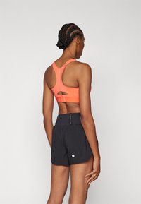 Koral sport-BH med racerback-design og udskæringsdetalje, parret med sorte shorts med reflekterende detaljer og en rund kant.
