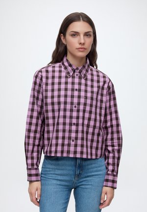 Femme portant une chemise à carreaux violette et noire à boutons, manches longues, et un jean bleu sur un fond blanc uni.