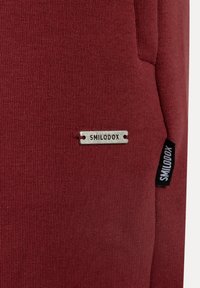 Sudadera de tela roja con superficie texturizada, una etiqueta de metal con la inscripción "SMILODOX" y una etiqueta de tela negra vertical en el lado.