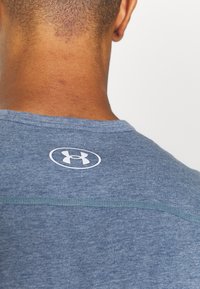 Camiseta deportiva de tela azul jaspeado con cuello redondo, que presenta un logo blanco en la parte posterior y detalles de costura en contraste.