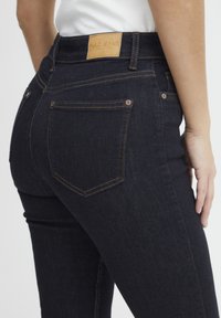 Sötétkék, slim fit farmernadrág, bőr derékkal, narancssárga varrással, két hátsó zsebbel és sima felülettel.