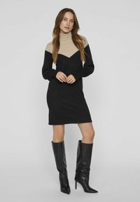 Robe en tricot avec un col montant, composée d'une partie supérieure beige et d'une jupe noire. Manches longues avec des poignets texturés. Associée à des bottes noires montant jusqu'aux genoux.