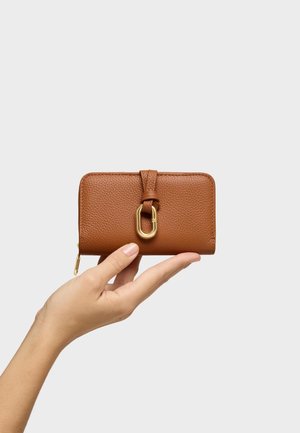 Dłoń trzymająca brązowy skórzany portfel z złotym metalowym zapięciem i zamkiem błyskawicznym na jednolitym jasnoszarym tle.