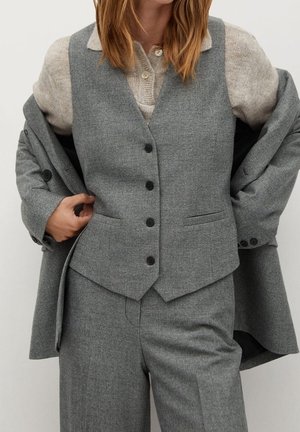 Frau trägt einen grauen Dreiteiler-Anzug und beige Strickpullover, richtet die Anzugjacke über ihren Schultern.