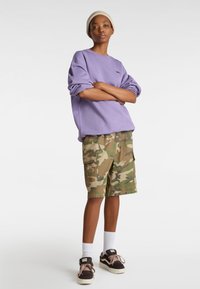 Sudadera extra grande morada, pantalones cortos cargo de camuflaje con múltiples bolsillos, calcetines blancos hasta la altura del tobillo y zapatillas Vans de color marrón oscuro.