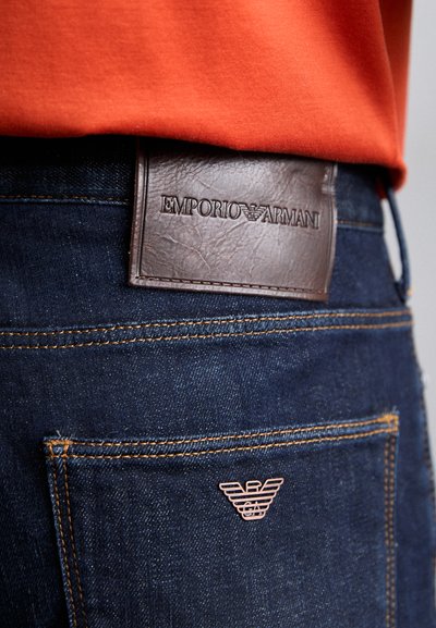 Dark blue denim jeans featuring a brown leather logo patch labeled "EMPORIO ARMANI" and a small metal emblem on the back pocket.