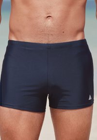 Mannelijke torso met donkerblauwe aansluitende zwembroek, staand tegen een wazige strandachtergrond.