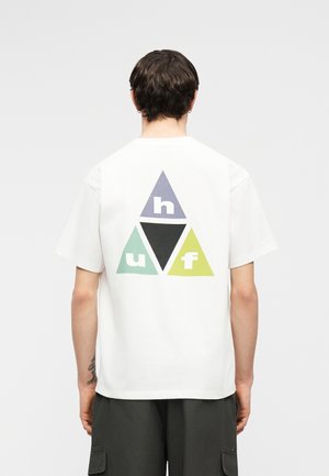 Persona con cabello corto y oscuro, vistiendo una camiseta blanca que presenta cuatro triángulos de colores con las letras "h", "u", "f" y un triángulo negro central en la parte trasera.