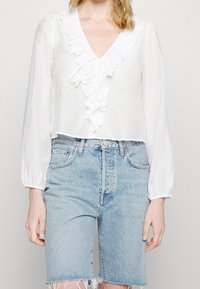 Blouse blanche avec décolleté en V à volants et manches longues froncées, associée à un short en jean bleu clair, avec ourlet effiloché et taille haute.