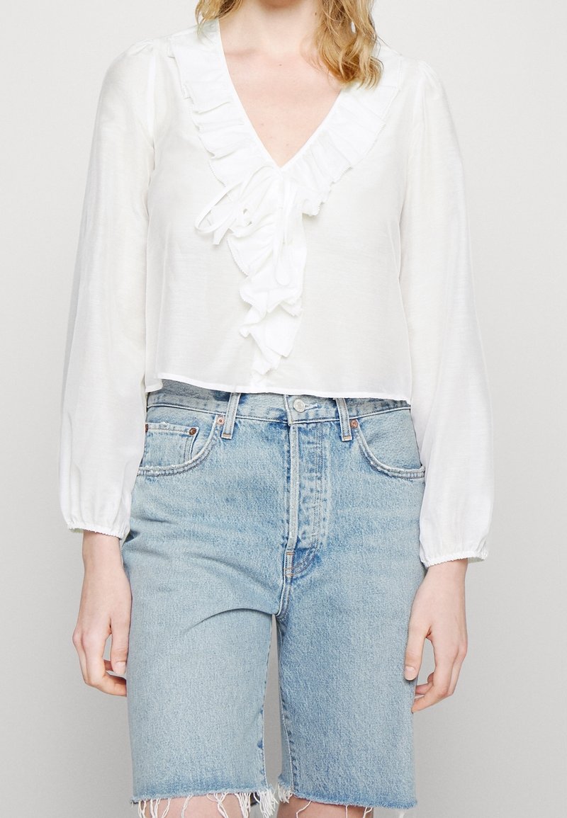Blouse blanche avec décolleté en V à volants et manches longues froncées, associée à un short en jean bleu clair, avec ourlet effiloché et taille haute.