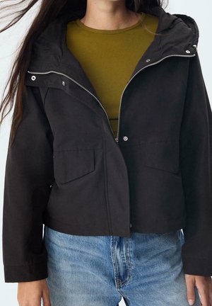 Personne portant une veste noire zippée avec des poches à rabat sur un haut vert olive et un jean bleu clair.