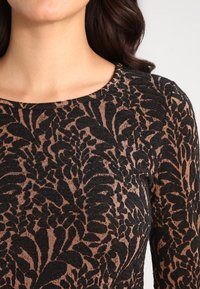 Långärmad topp med rundad halsringning som har ett svart blommigt mönster på en beige bakgrund, tillverkad av ett texturerat, stickat tyg.