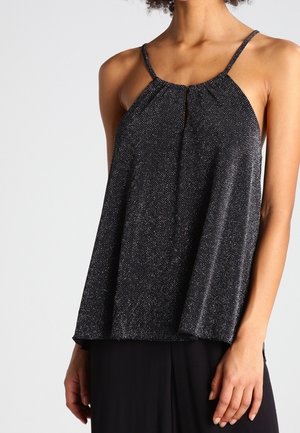 Kobieta ubrana w czarny top na szyję bez rękawów z subtelnym wzorem diamentowym, w połączeniu z czarnymi spodniami, na jednolitym tle.