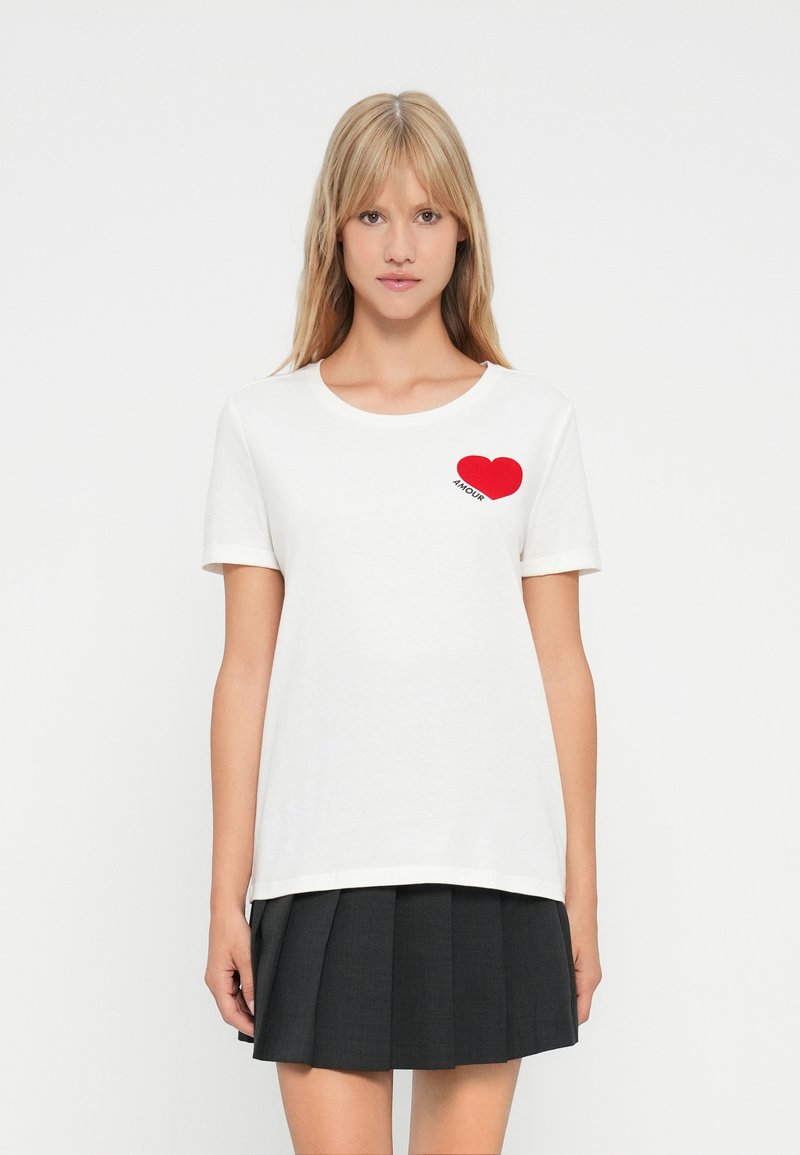 Ξανθιά γυναίκα φορώντας λευκό μπλουζάκι με κόκκινη καρδιά και κείμενο "AMOUR", σε συνδυασμό με μαύρη πλισέ μίνι φούστα, στέκεται μπροστά σε λευκό φόντο.