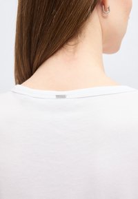 Dos d'une femme avec des cheveux bruns raides, portant une chemise blanche avec une petite étiquette argentée près de l'encolure et une boucle d'oreille argentée.