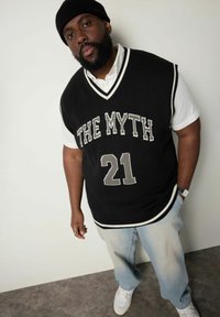 Schwarzer gestrickter Weste mit "THE MYTH" und "21" in Grau, weiß gestreiftem Saum, über einem weißen Hemd mit Kragen getragen, kombiniert mit hellen Jeans.