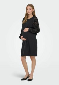 Robe de maternité en jersey noir avec des manches longues couvertes de dentelle. Design à hauteur du genou, coupe décontractée, avec col rond et poignets côtelés.