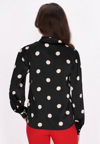 Blusa nera con grandi pois beige, caratterizzata da maniche lunghe e polsini con bottoni. Tessuto morbido e leggero con colletto classico.