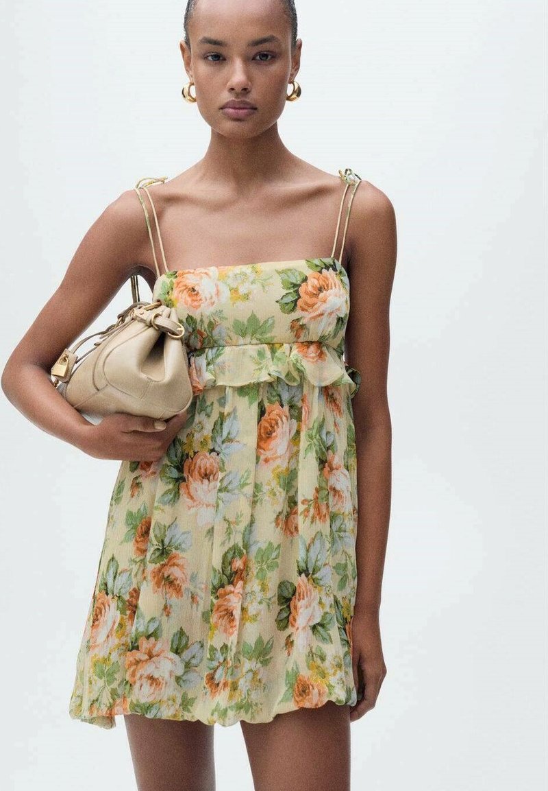 Blumenkleid in hellgelb mit tailliertem Oberteil, rüschigem Saum und verstellbaren Spaghetti-Trägern. Das Model hält eine beige Tasche.