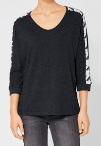 Kvinna klädd i en mörkgrå långärmad tröja med vita mönstrade ränder på ärmarna och mörkgrå jeans, stående med avslappnade armar.