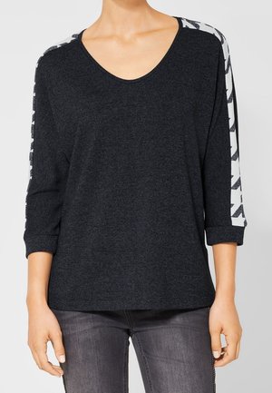 Vrouw in een donkergrijze lange mouwen shirt met witte gestreepte patronen op de mouwen en donkergrijze jeans, staande met ontspannen armen.