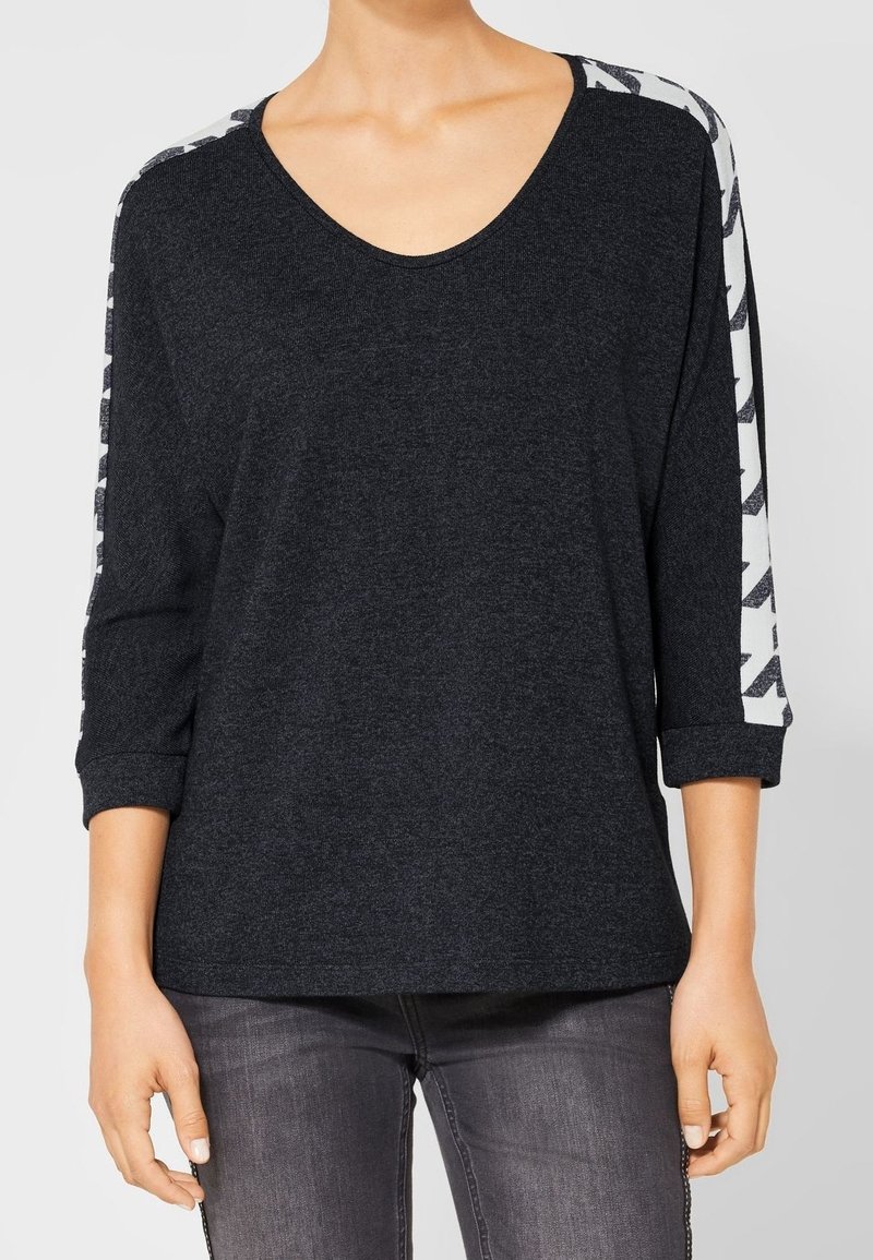 Kvinna klädd i en mörkgrå långärmad tröja med vita mönstrade ränder på ärmarna och mörkgrå jeans, stående med avslappnade armar.