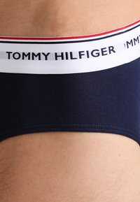 Marineblaue Baumwollslips mit weichem Bund, der von einem weißen und roten gestreiften Rand umgeben ist, und geprägtem "TOMMY HILFIGER" Logo.