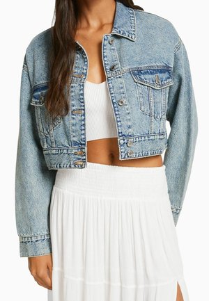Femme portant une veste en jean bleu clair courte sur un crop top blanc côtelé et une jupe plissée blanche avec une fente latérale.
