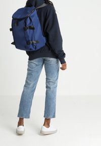Osoba ubrana w niebieskie dżinsy, białe trampki, granatowy hoodie i nosząca dużą niebieską plecak, stojąca na białej podłodze na białym tle.