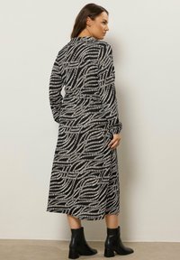 Kobieta w czarno-białej, wzorzystej, midi sukience z długim rękawem i czarnych botkach za kostkę, stojąca na tle gładkiej, jasnej ściany.