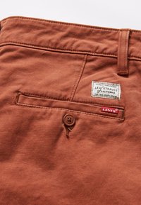 Poche arrière d'un pantalon Levi's couleur rouille montrant une fermeture par bouton, une boucle de ceinture et deux étiquettes de marque, dont une étiquette rouge Levi's et un patch en tissu cousu.