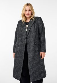 Cappotto grigio a spina di pesce con chiusura doppiopetto, rever a dog legato e due tasche laterali. Realizzato in tessuto testurizzato, arriva fino a metà coscia.