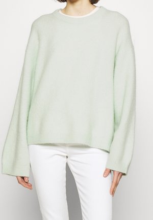 Osoba ubrana w luźny, jasnozielony sweter z długimi rękawami i białe spodnie, stojąca na tle jednolitego tła.