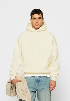 Homme portant un sweat à capuche écru et un jean bleu, tenant un pull tricoté beige dans sa main gauche, sur un fond blanc uni.