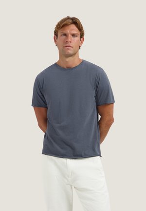 Man met lichtbruin haar, gekleed in een effen donkerblauw T-shirt met korte mouwen en witte broek, staand met handen achter de rug.