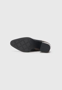 Semelle d'une chaussure habillée à bout pointu avec une semelle extérieure en caoutchouc noire à motifs et un petit talon bloc sur un fond clair uni.