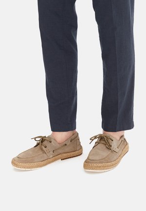 Person trägt beige Wildleder-Schnür-Espadrilles mit geflochtenen Jutésohlen und dunkelblauen, knöchellangen Hosen.