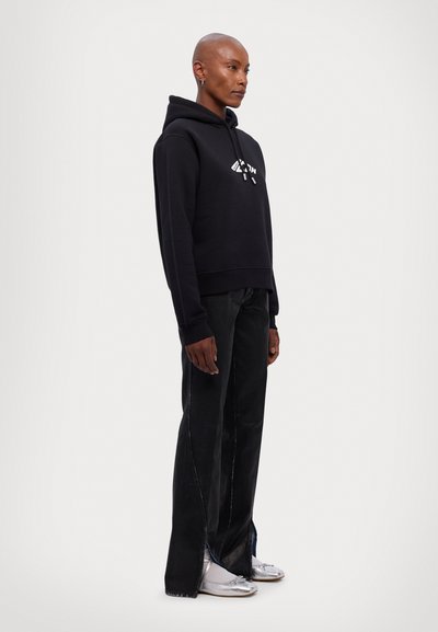 Schwarzer Kapuzenpullover mit einem Frontlogo, kombiniert mit lockeren schwarzen Hosen mit seitlichen Schlitzen. Das Model trägt silberne Schuhe.