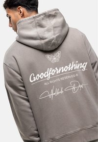 Haut à capuche gris avec une capuche, un détail de papillon brodé, et un texte blanc indiquant "Goodfornothing" et "Worldwide Dept" au dos.