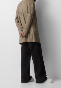 Persona vestida con una chaqueta de ante beige, pantalones de pierna ancha negros y zapatillas blancas, de pie con las manos entrelazadas detrás de la espalda contra una pared blanca.