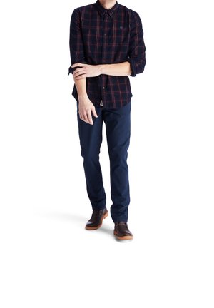 Mann, der seine hochgekrempelten Ärmel anpasst, trägt ein dunkel kariertes Hemd, marineblaue Hose und braune Lederschuhe und steht vor weißem Hintergrund.