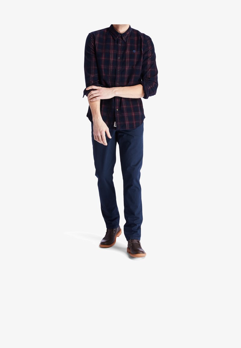Homme ajustant ses manches retroussées, portant une chemise à carreaux sombre, un pantalon bleu marine et des chaussures en cuir marron, debout sur un fond blanc.