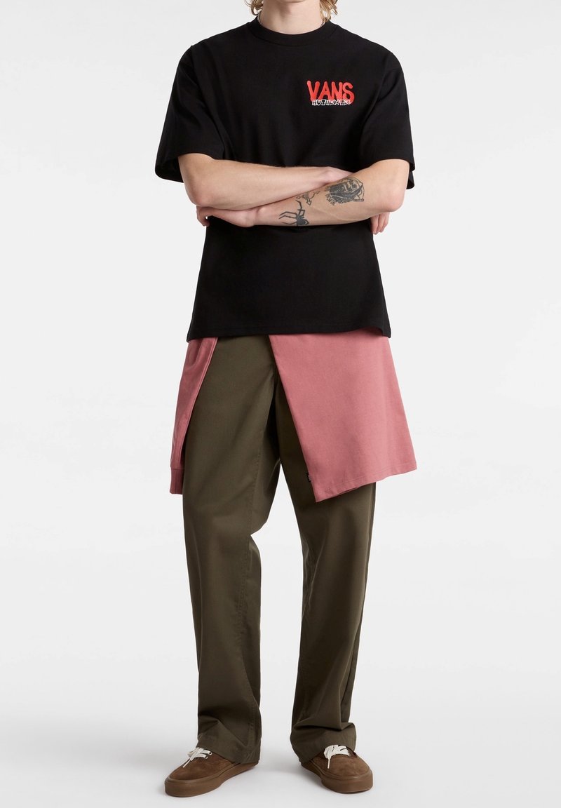 Personne portant un t-shirt Vans noir, un sweat rose noué à la taille, un pantalon vert olive et des chaussures marron, debout les bras croisés.