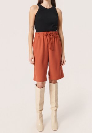 Donna che indossa una canottiera nera, pantaloni corti color ruggine fino al ginocchio con coulisse e stivali color crema fino al ginocchio, in piedi contro uno sfondo neutro.