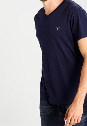 Mężczyzna noszący granatową koszulkę GANT z dekoltem w serek i ciemnoniebieskie dżinsy na jasnym, jednolitym tle.