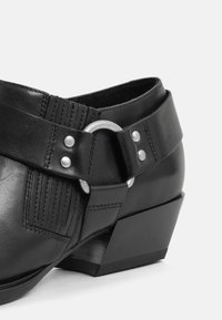 Scarpa nera in pelle con una fascia testurizzata, fibbia circolare in argento e borchie decorative. Presenta un tacco basso a blocco e cuciture ben definite.