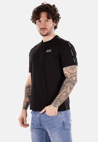 Camiseta corta de manga negra con un parche de marca en el pecho y acentos decorativos en los hombros, hecha de tela de algodón.