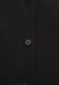 Černý vlněný kabát s jemnou texturou, který má dva knoflíky a rovný střih. Jednoduchý, minimalistický styl bez viditelných vzorů.